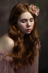 Fototapeta premium Young woman with long red curly hair in pink fairy dress with open shoulders and puff sleeves with pink flower in hair. Romantic portrait of girl on brown background