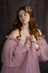 Young woman with long red curly hair in pink fairy dress with open shoulders and puff sleeves with pink flower in hair. Romantic portrait of girl on brown background