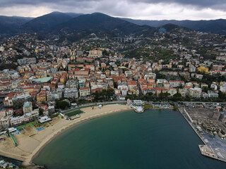 Fototapeta na wymiar Aerial view of Sanremo, Italian city on the seashore in Liguria, north Italy. Drone flying along the port over beaches and boardwalk with palm trees and Birds Eye of yacht parking in San Remo, Italy.
