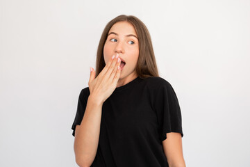 Portrait of flirty young woman covering mouth with hand over white background. Caucasian lady wearing black T-shirt looking away and yawning. Amazement concept