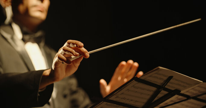 Male orchestra conductor controlling music in orchestra pit by movement of his hands and white baton, studio shot on black background 