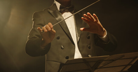 Male orchestra conductor wearing tux standing in front of music stand, controlling musicians by...