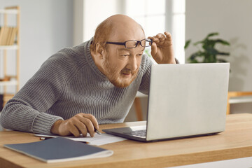 Eyesight problems. Confused mature man who has poor eyesight and works on laptop at home wearing...