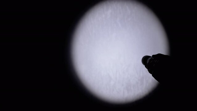 Unrecognizable Person Shines a Flashlight on the Wall in a Dark Room. Close-up. Silhouette of a hand with a flashlight illuminates the wall in dark. Spot or beam of light from a lantern. Shadow.