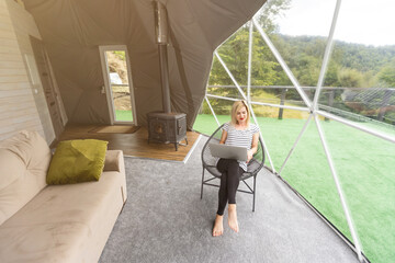 Happy young woman chatting online by using laptop in dome camping. Glamping vacation concept