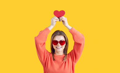 Happy young woman wishes you to have fun and find love on Valentine's Day. Pretty girl in funny heart shaped glasses standing isolated on yellow background, smiling and holding up red Valentine card