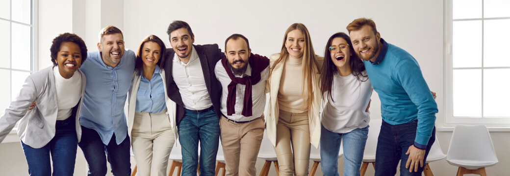 Diverse team of happy united business people hugging each other in the office. Mixed race group of young men and women standing in row, huddling, smiling and laughing together. Unity, teamwork concept