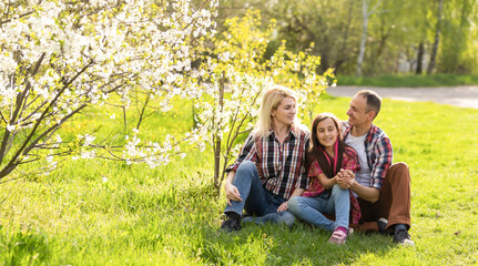 Fototapeta premium Outdoor portrait of happy young family playing in spring park under blooming tree, lovely couple with little child having fun in sunny garden