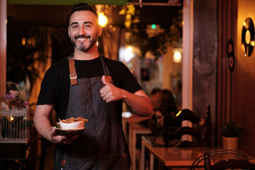Waiter showing thumb up and smiling while holding a plate with food in a restaurant.