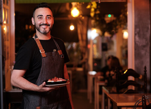Waiter in uniform smiling at camera while holding a plate with food in a restaurant.