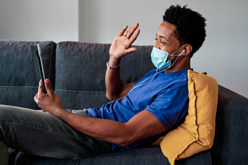 Man with face mask having a video call with digital tablet while lying on sofa at home.