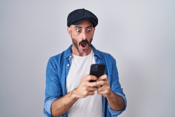 Hispanic man with beard using smartphone typing message in shock face, looking skeptical and sarcastic, surprised with open mouth