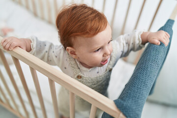Adorable redhead toddler standing on cradle with relaxed expression at bedroom