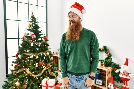 Redhead man with long beard wearing christmas hat by christmas tree winking looking at the camera with sexy expression, cheerful and happy face.