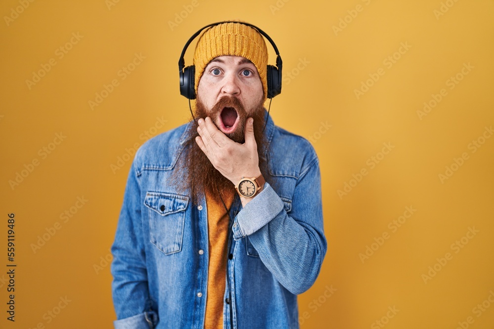 Wall mural caucasian man with long beard listening to music using headphones looking fascinated with disbelief,