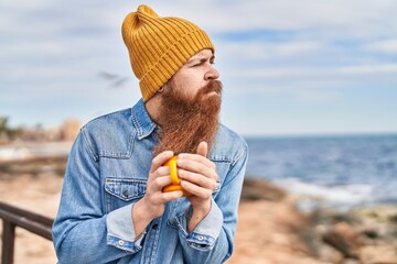 Young redhead man drinking cup of coffee with serious expression at seaside