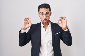 Handsome business hispanic man standing over white background looking surprised and shocked doing ok approval symbol with fingers. crazy expression