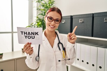 Young caucasian woman wearing doctor uniform holding thank you banner smiling happy pointing with hand and finger to the side