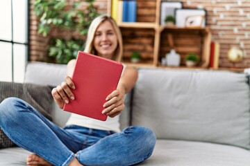 Obraz premium Young blonde woman holding book sitting on sofa at home