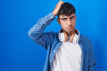 Young hispanic man standing over blue background confuse and wondering about question. uncertain with doubt, thinking with hand on head. pensive concept.