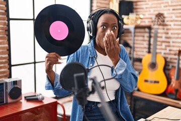 Beautiful black woman holding vinyl record at music studio covering mouth with hand, shocked and afraid for mistake. surprised expression