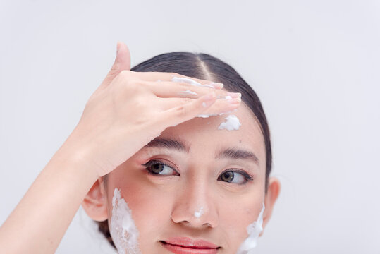 Beautiful lady applying facial wash on her forehead. Asian woman scrubbing face with an exfoliating soap. Studio photo with plain white background.