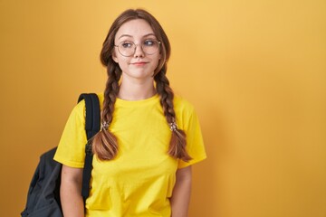 Young caucasian woman wearing student backpack over yellow background smiling looking to the side and staring away thinking.