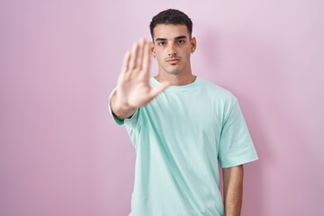 Handsome hispanic man standing over pink background doing stop sing with palm of the hand. warning...