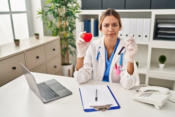 Young blonde woman wearing doctor uniform holding blood and heart skeptic and nervous, frowning...