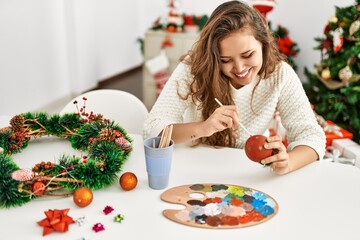 Young beautiful hispanic woman painting christmas ball sitting on table at home