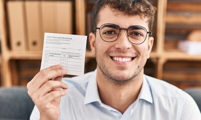 Young hispanic man holding covid record card looking positive and happy standing and smiling with a confident smile showing teeth