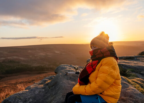 Woman in yellow coat and orange scarf reaching the destination and sitting   on top of hill or mountain and recording video  at sunrise. British cold winter. Local tourism concept.