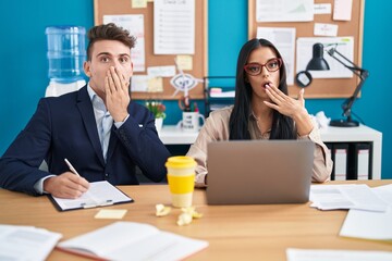 Young hispanic man and woman working at the office covering mouth with hand, shocked and afraid for...