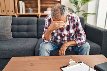 Senior grey-haired man patient stressed with hands on face sitting on sofa at psychology clinic