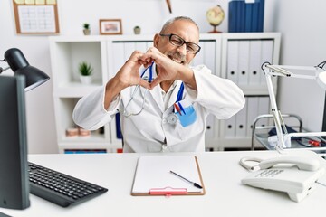 Mature doctor man at the clinic smiling in love doing heart symbol shape with hands. romantic concept.