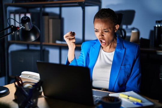Beautiful african american woman working at the office at night angry and mad raising fist frustrated and furious while shouting with anger. rage and aggressive concept.