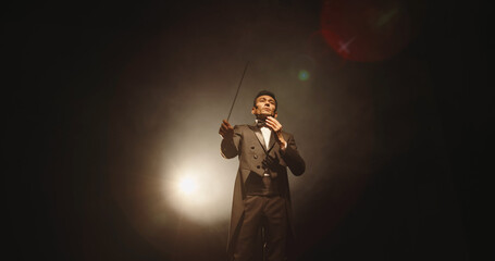 Asian male orchestra conductor wearing tux is directing symphony orchestra with movement of his hands and stick, studio shot on black background  © andreybiling
