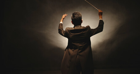 Unrecognizable Male orchestra conductor controlling music in orchestra pit by movement of his hands and white baton, studio shot on black background  - obrazy, fototapety, plakaty