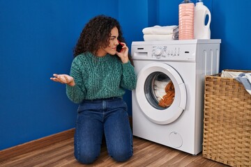 Young beautiful hispanic woman talking on smartphone with technician for broke washing machine at laundry room