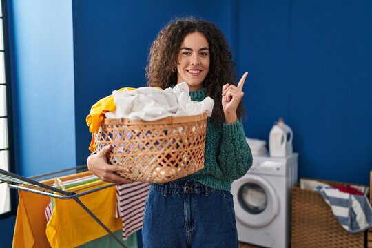 Young hispanic woman holding laundry basket smiling happy pointing with hand and finger to the side