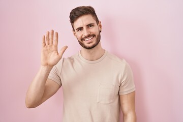 Hispanic man with beard standing over pink background waiving saying hello happy and smiling, friendly welcome gesture