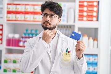 Hispanic man with beard working at pharmacy drugstore holding credit card serious face thinking about question with hand on chin, thoughtful about confusing idea