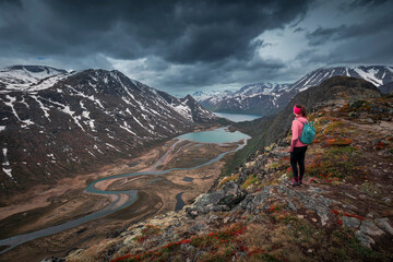 Girl hiking along turquoise lakes and river in mountain landscape from above the hike to Knutshoe summit in Jotunheimen National Park in Norway, snow-covered mountains of Besseggen in background