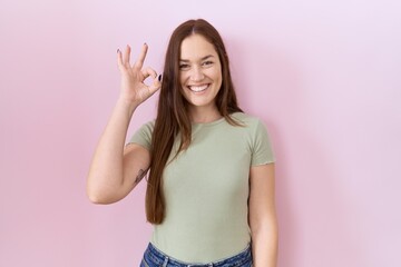 Beautiful brunette woman standing over pink background smiling positive doing ok sign with hand and fingers. successful expression.