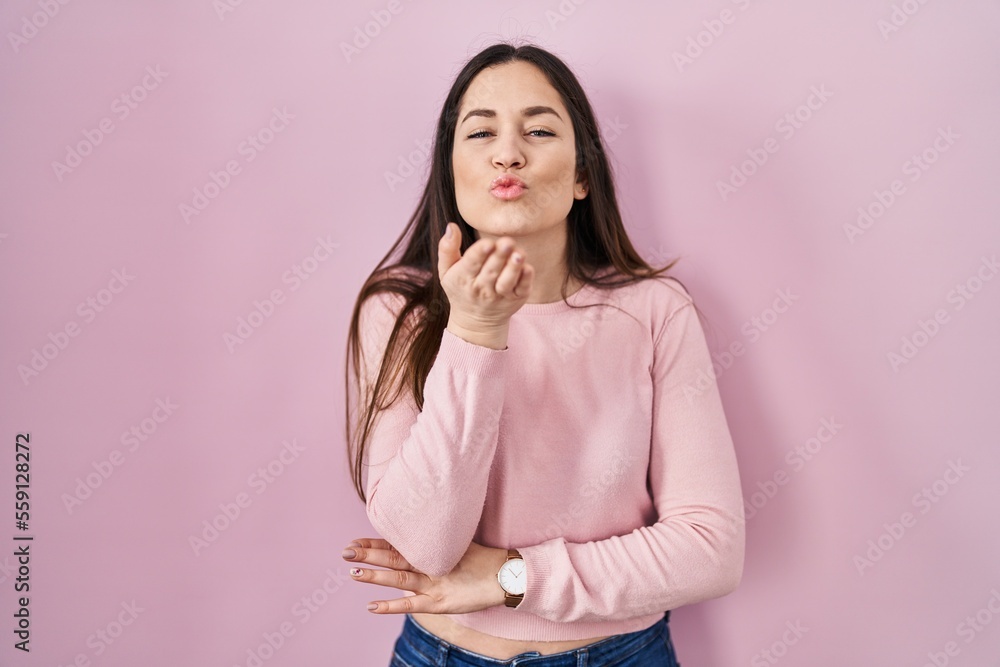 Wall mural Young brunette woman standing over pink background looking at the camera blowing a kiss with hand on air being lovely and sexy. love expression.