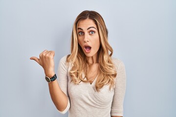 Fototapeta na wymiar Young blonde woman standing over isolated background surprised pointing with hand finger to the side, open mouth amazed expression.