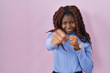 African woman standing over pink background punching fist to fight, aggressive and angry attack,...