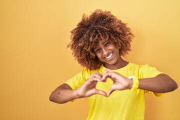 Young hispanic woman with curly hair standing over yellow background smiling in love doing heart symbol shape with hands. romantic concept.