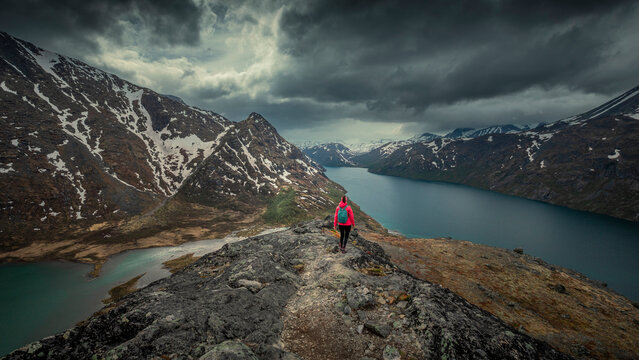 Woman hiking along blue lake in mountain landscape of Knutshoe in Jotunheimen National Park in Norway, mountains of Besseggen in background, dramatic cloudy sky