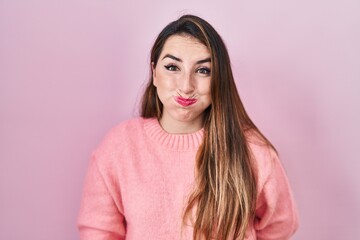 Fototapeta premium Young hispanic woman standing over pink background puffing cheeks with funny face. mouth inflated with air, crazy expression.